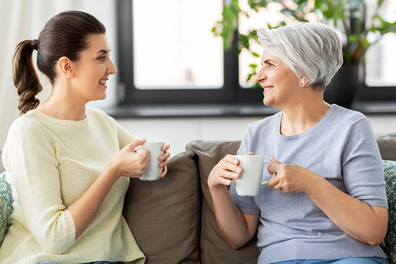 family, generation and people concept - happy smiling senior mother with adult daughter drinking coffee or tea and talking at home